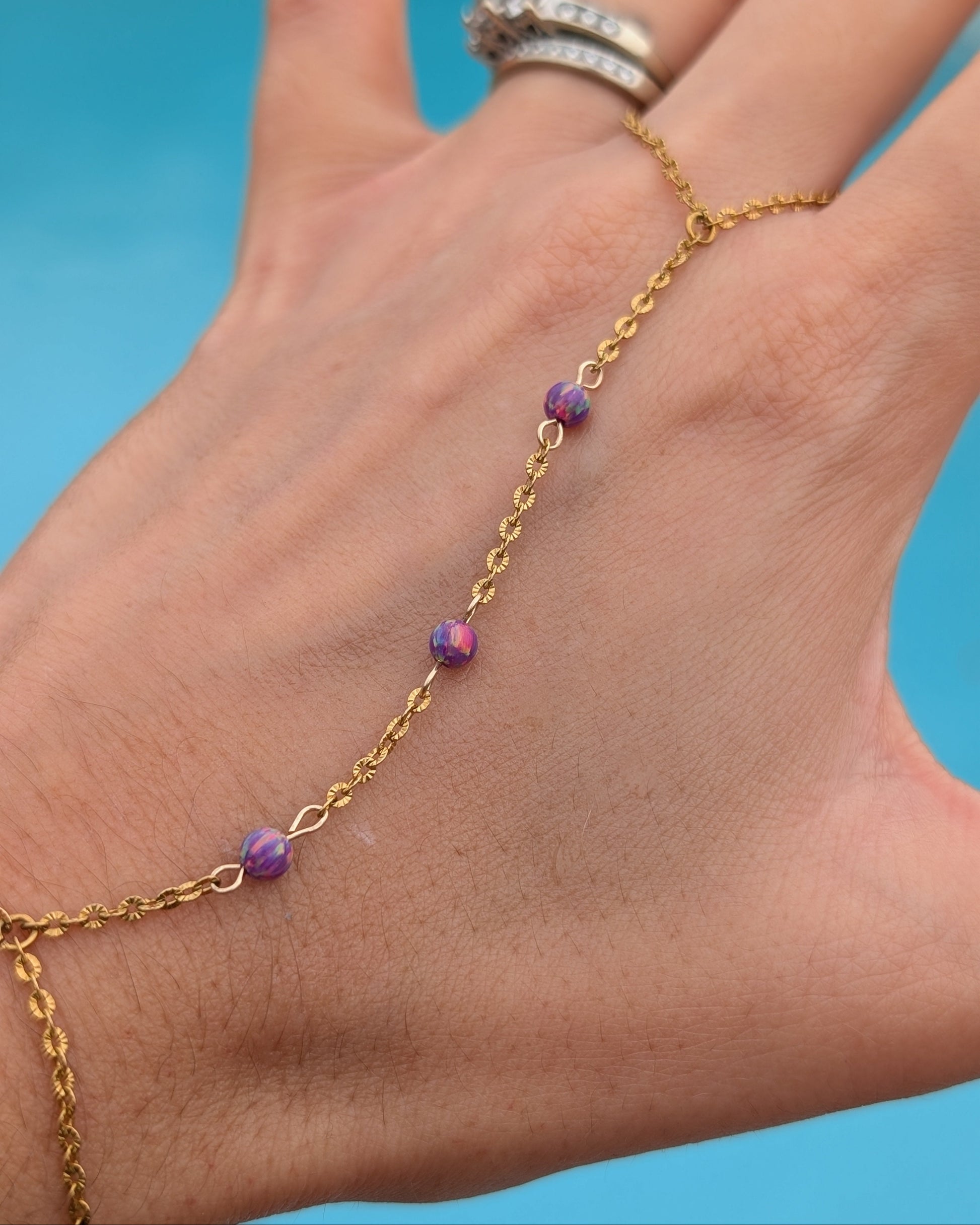 A gold chain hand bracelet with three small purple and pink iridescent beads, shown worn on a hand against a bright blue background.