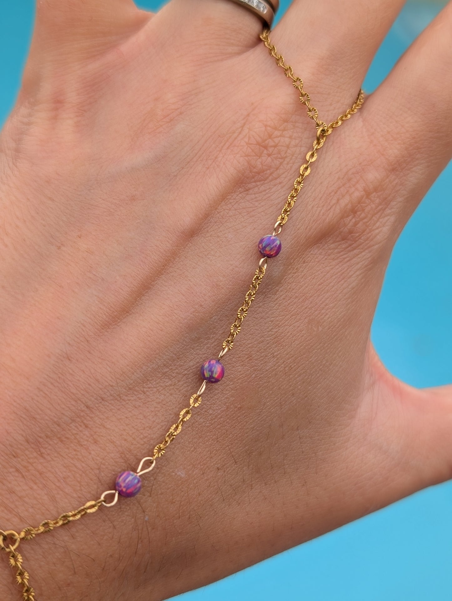 A gold chain anklet with three small purple marbled beads worn on a person's ankle, shown against a bright blue background.