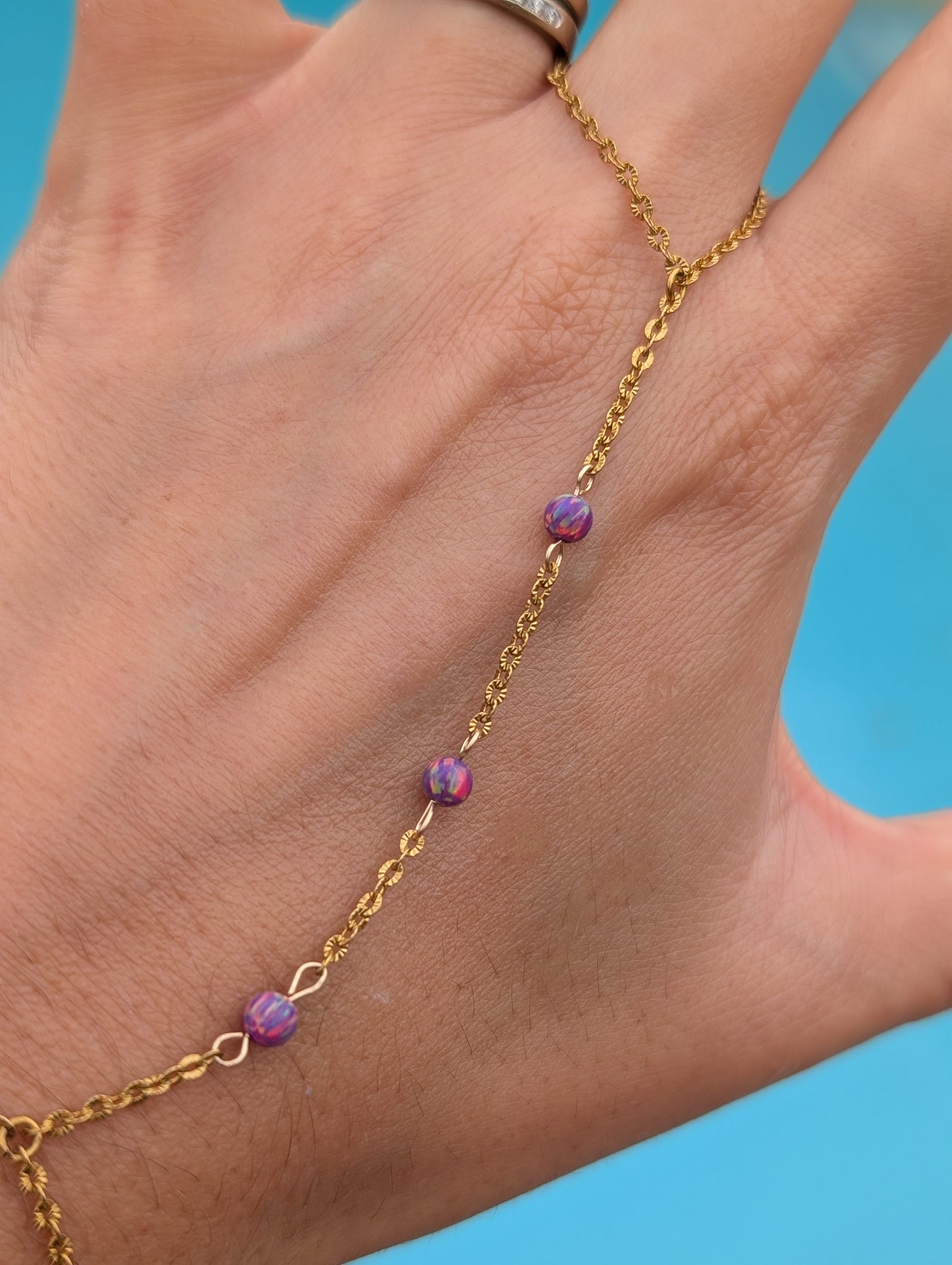 A gold chain anklet with three small purple marbled beads worn on a person's ankle, shown against a bright blue background.