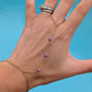 A gold chain hand chain bracelet with small purple beads, shown worn on a person's hand with matching rings on the index finger, set against a bright blue background.