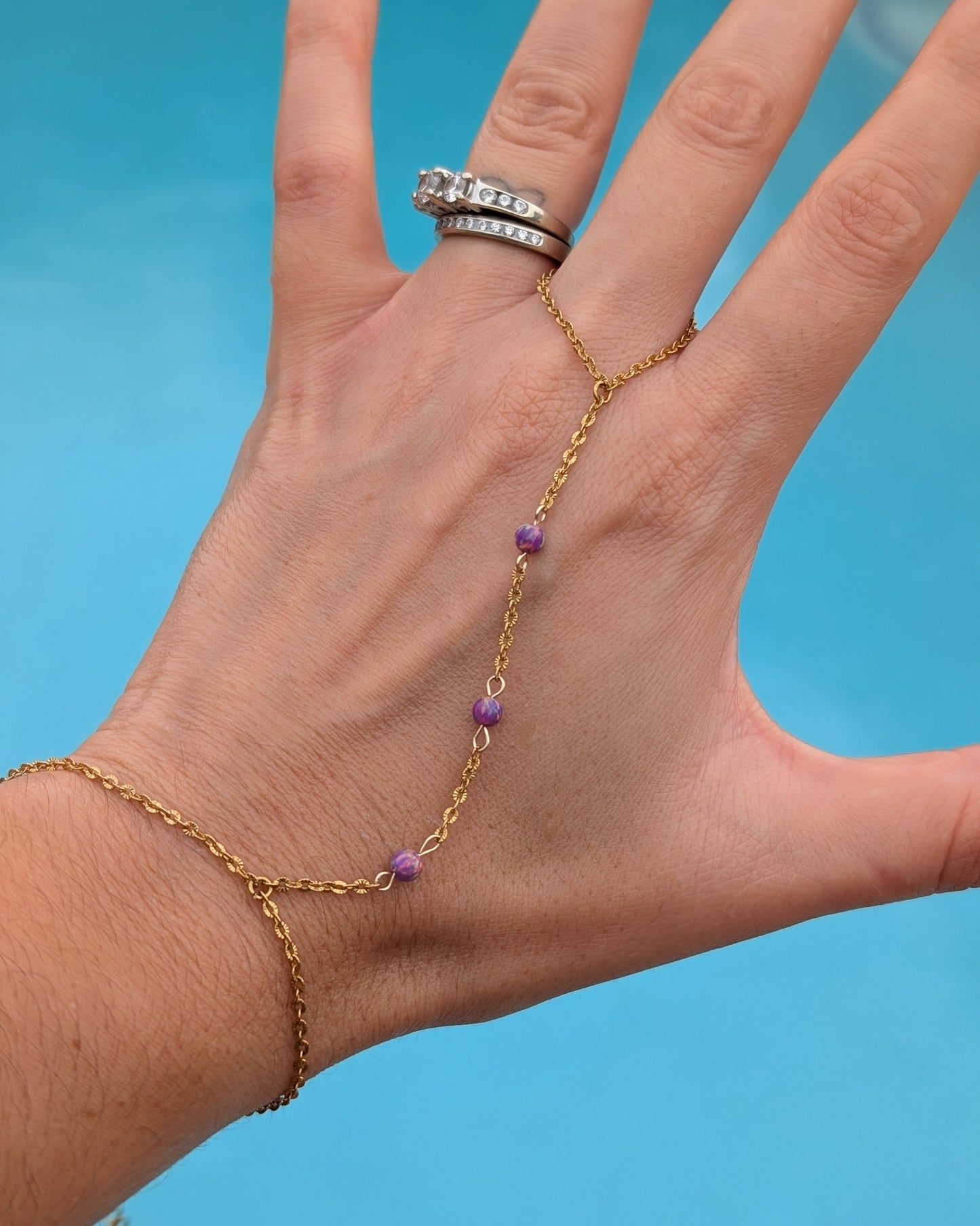A gold chain hand chain bracelet with small purple beads, shown worn on a person's hand with matching rings on the index finger, set against a bright blue background.