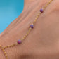 A gold chain anklet with three small purple marbled beads, shown worn on a person's ankle against a bright blue background.