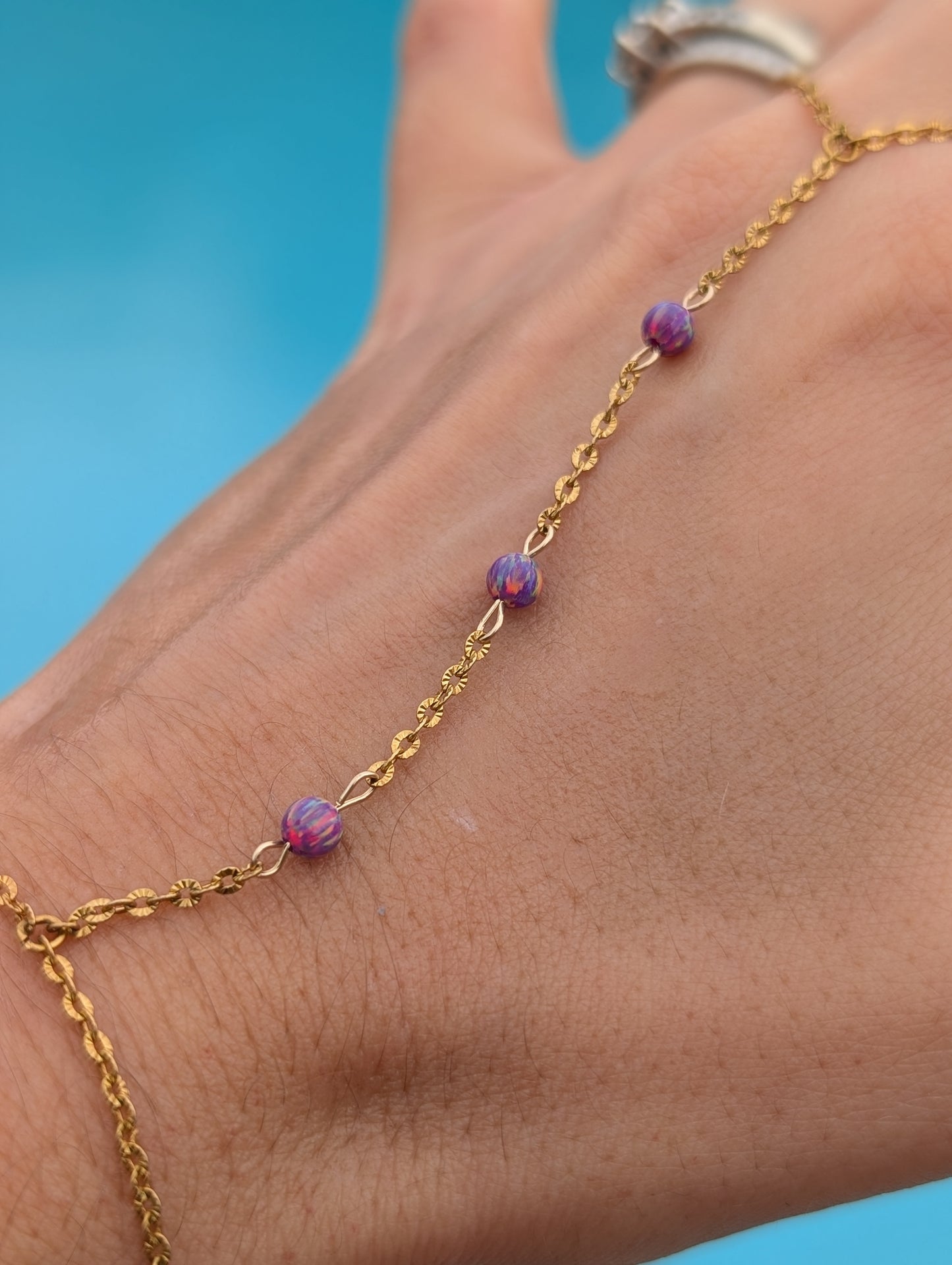 A gold chain anklet with three small purple marbled beads, shown worn on a person's ankle against a bright blue background.