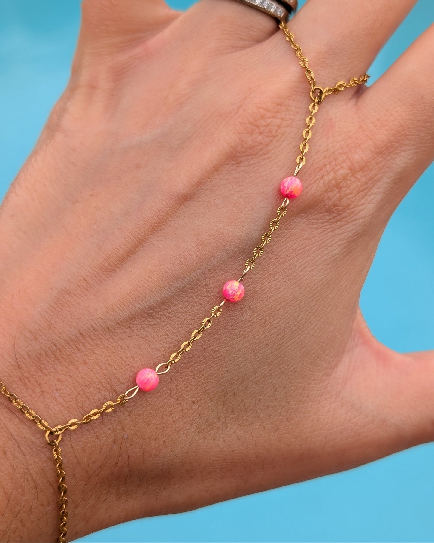 A gold chain hand and wrist bracelet with small pink beads, shown worn on a person's hand against a bright blue background.