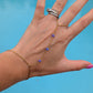 A gold chain hand and wrist bracelet with small purple beads, shown worn on a person's hand against a bright blue background.