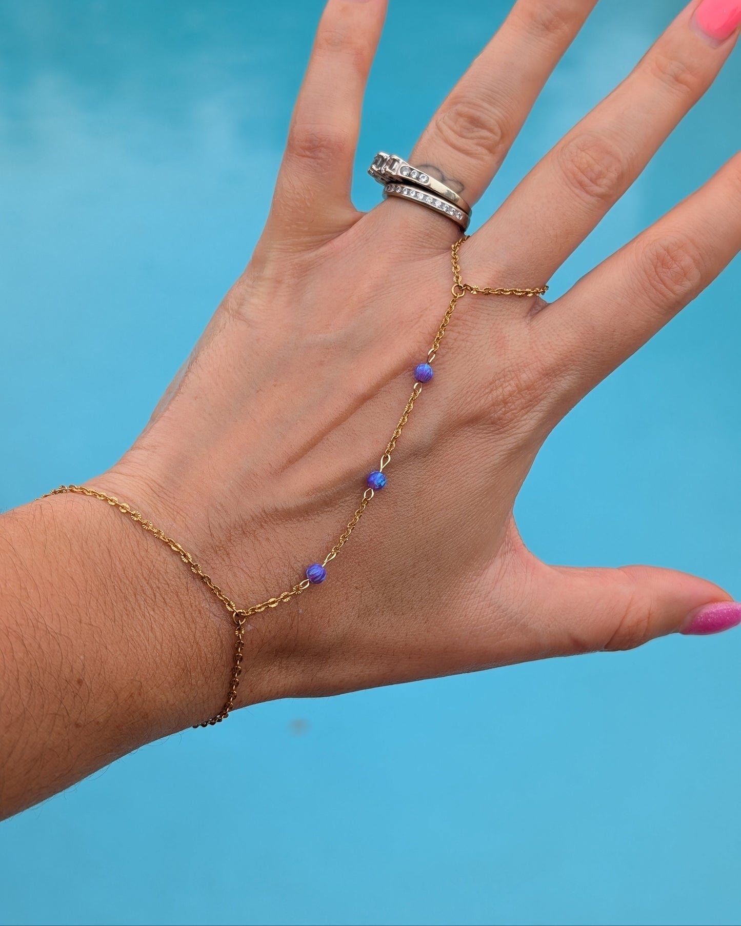 A gold chain hand and wrist bracelet with small purple beads, shown worn on a person's hand against a bright blue background.