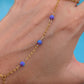 A gold chain anklet with three small purple and blue marbled beads worn on a person's ankle, shown against a bright blue background.