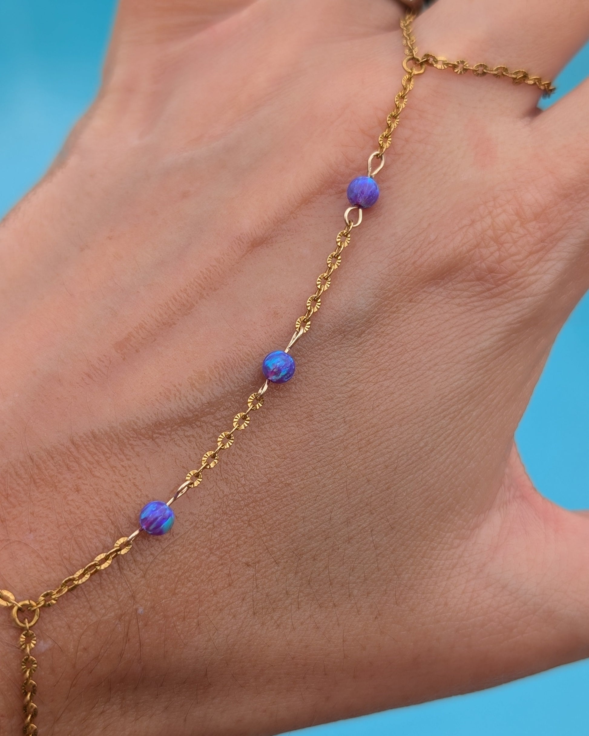 A gold chain anklet with three small purple and blue marbled beads worn on a person's ankle, shown against a bright blue background.
