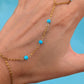 A delicate gold chain hand bracelet with four small turquoise blue beads, shown worn on a person's hand against a bright blue background.