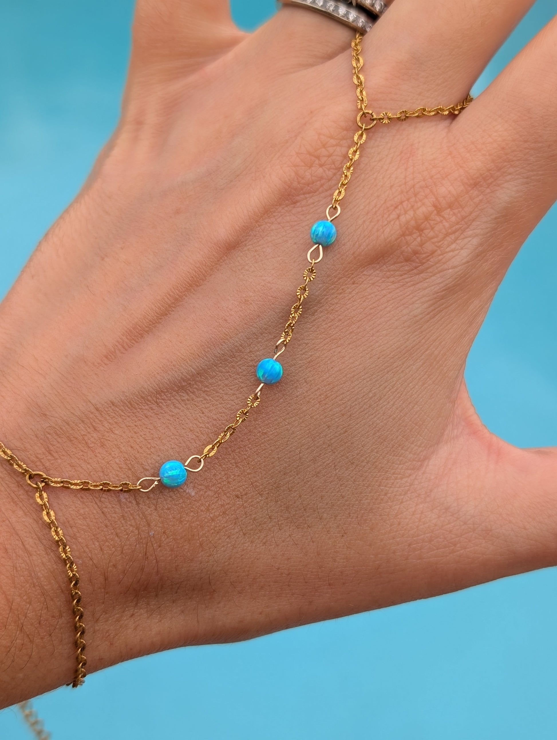 A delicate gold chain hand bracelet with four small turquoise blue beads, shown worn on a person's hand against a bright blue background.